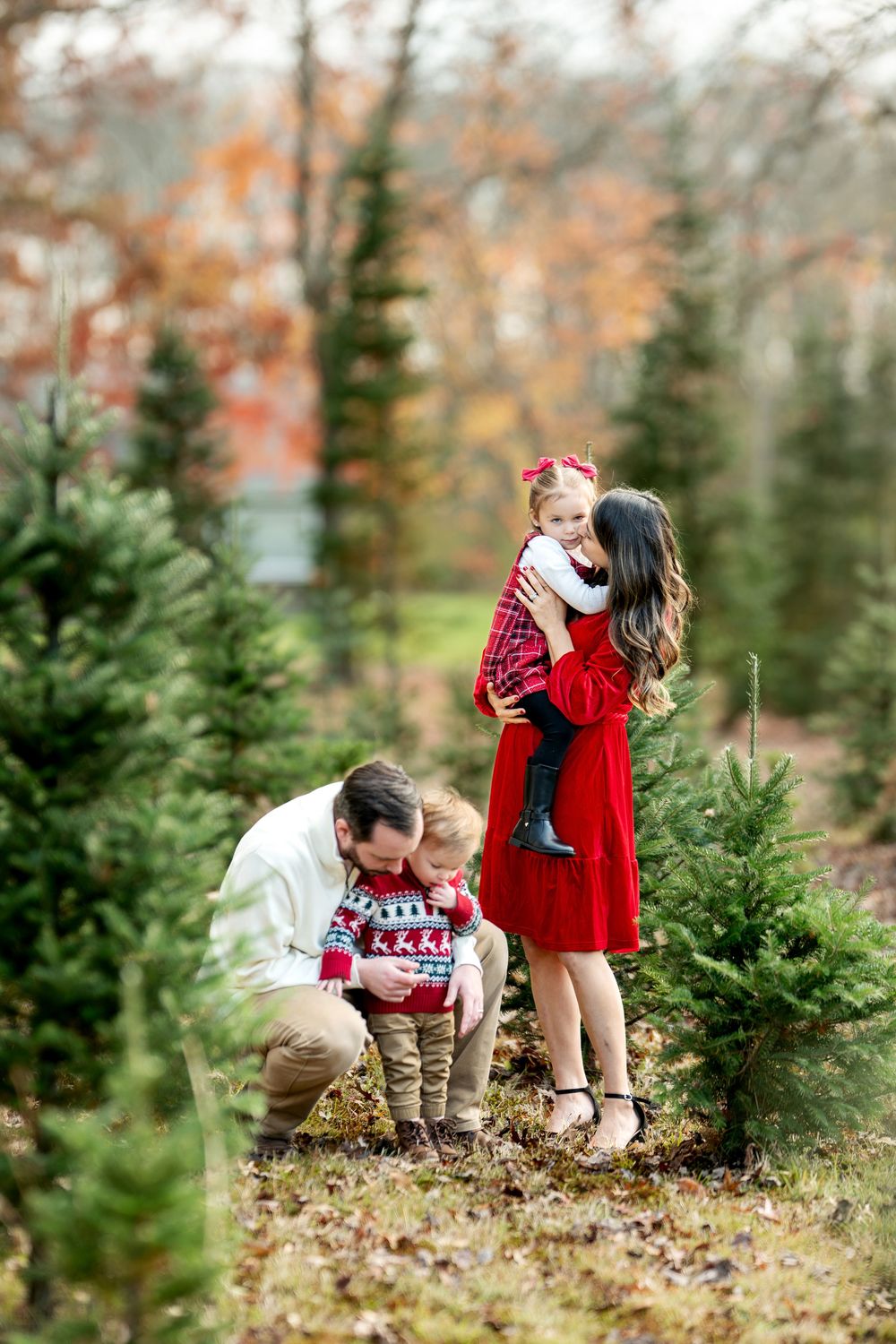 Family of four interacts warmly among Christmas trees, children dressed in festive red holiday clothing.