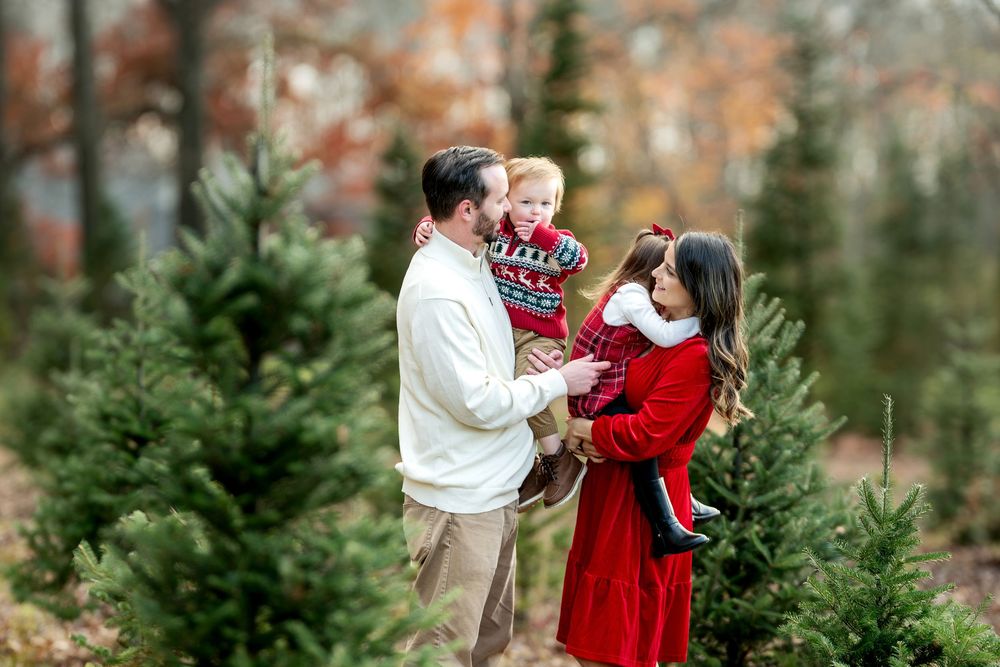 Family of four in red and white holiday outfits poses together among green Christmas trees outdoors.