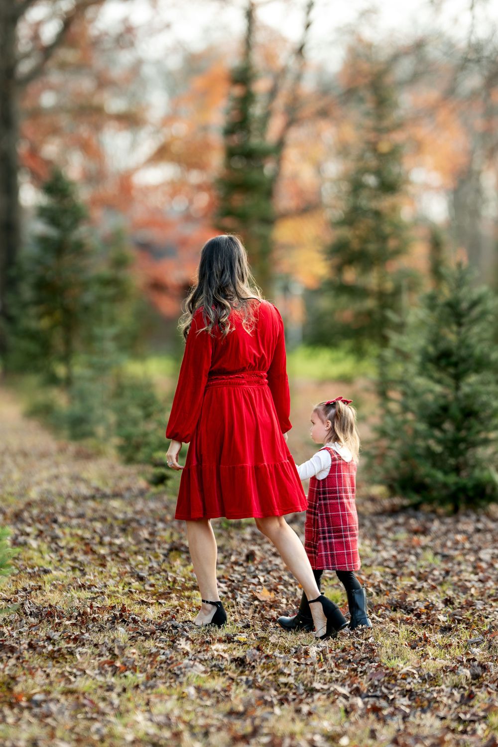 Mother in red dress and daughter in plaid dress walking hand in hand at a Christmas tree farm.