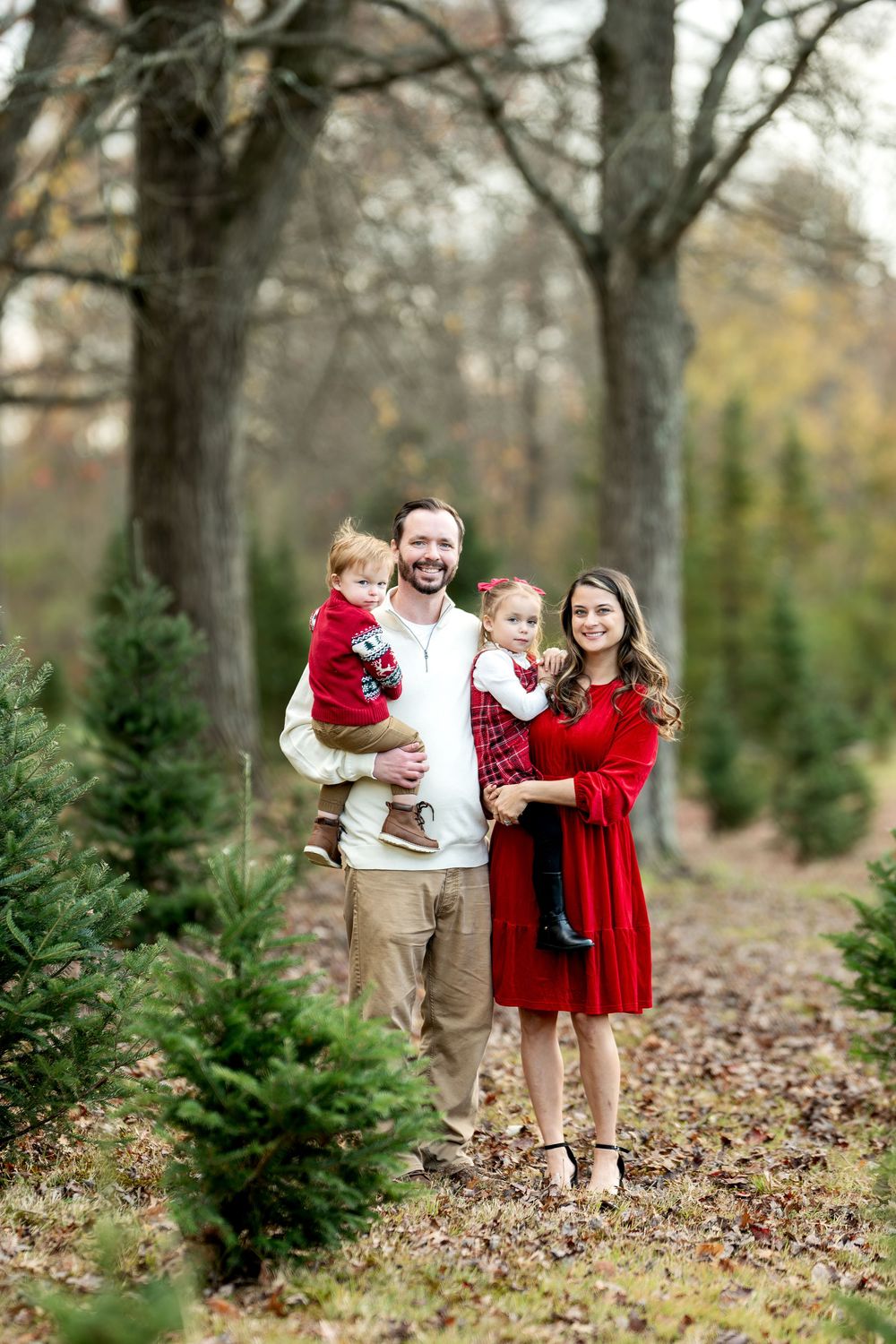 A smiling family of four stands among evergreen trees at a Christmas tree farm in autumn.