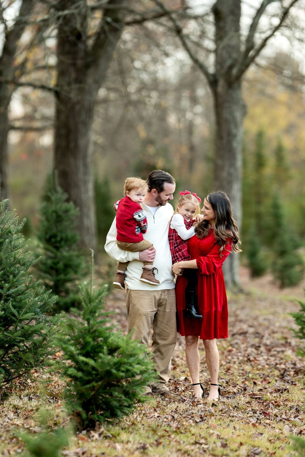 Family of four poses among Christmas trees, parents holding children in festive red holiday outfits outdoors.