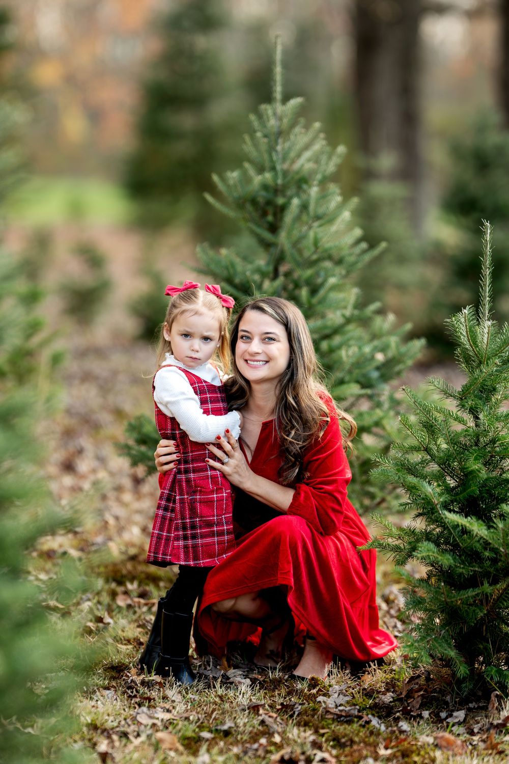 Mother in red dress kneels beside young daughter in plaid dress among Christmas trees, both smiling.