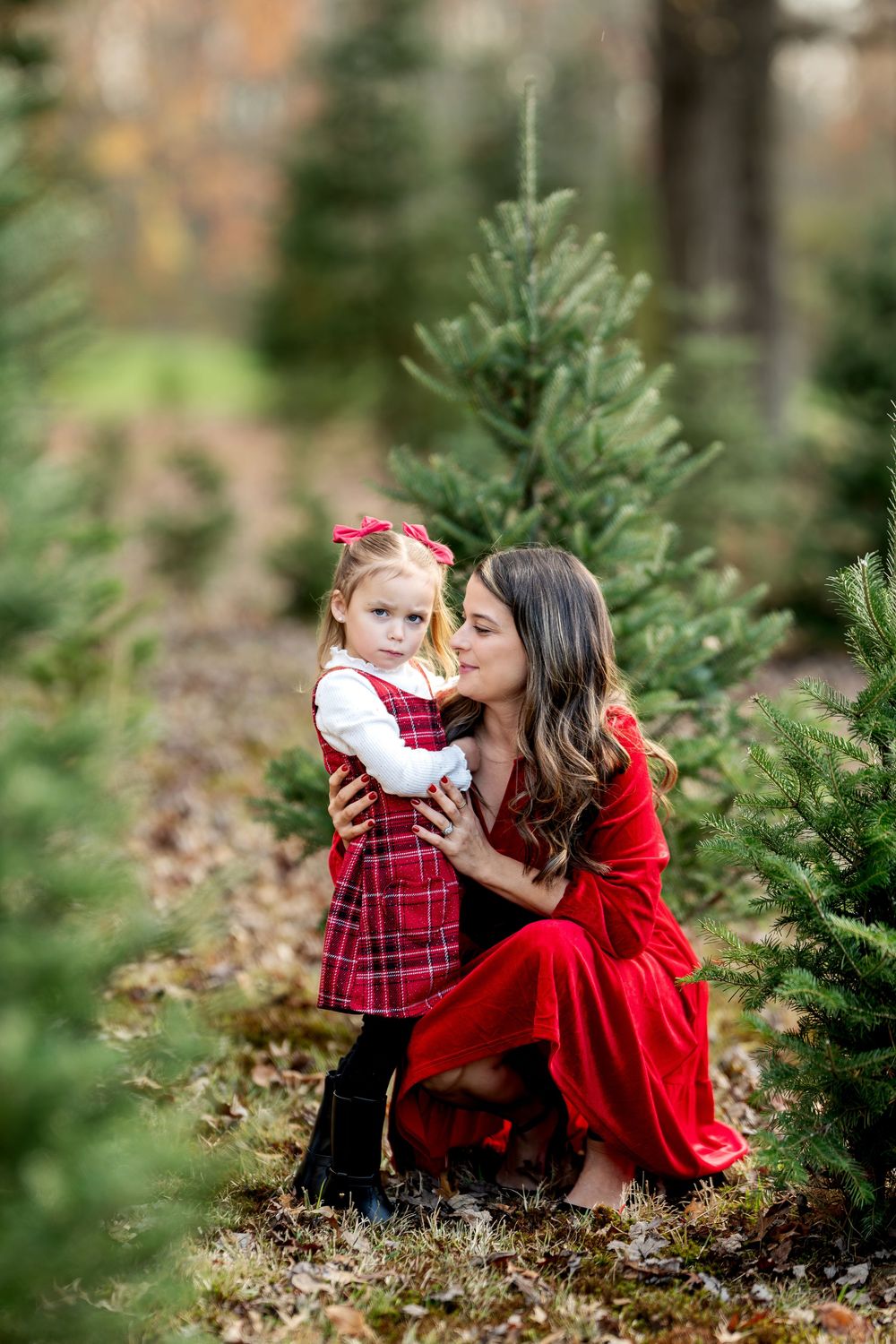 Mom in red dress tenderly kisses young daughter in plaid outfit while kneeling among Christmas trees.