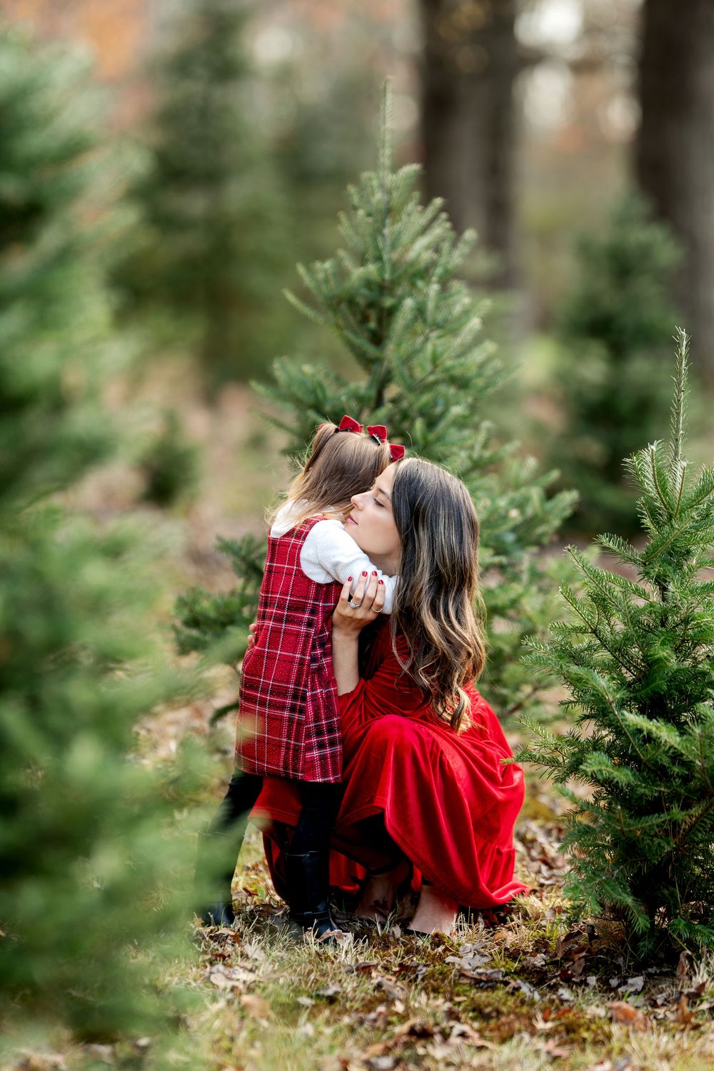 Sweet moment as young girl in plaid dress kisses her mother in red among lush Christmas trees.