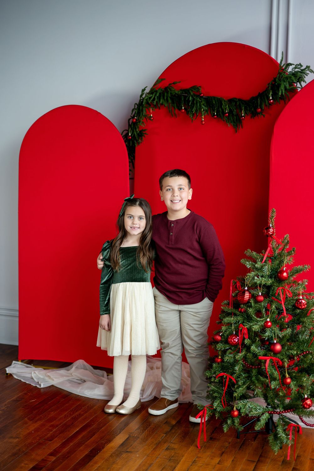 A smiling boy and girl in holiday clothes stand before red arch panels and a decorated Christmas tree.
