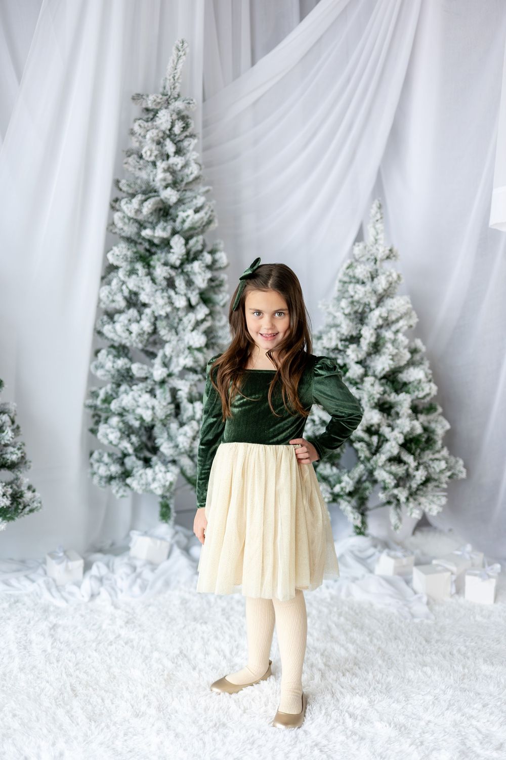 A girl in a green velvet top and cream tulle skirt poses sassily among snow-flocked Christmas trees.