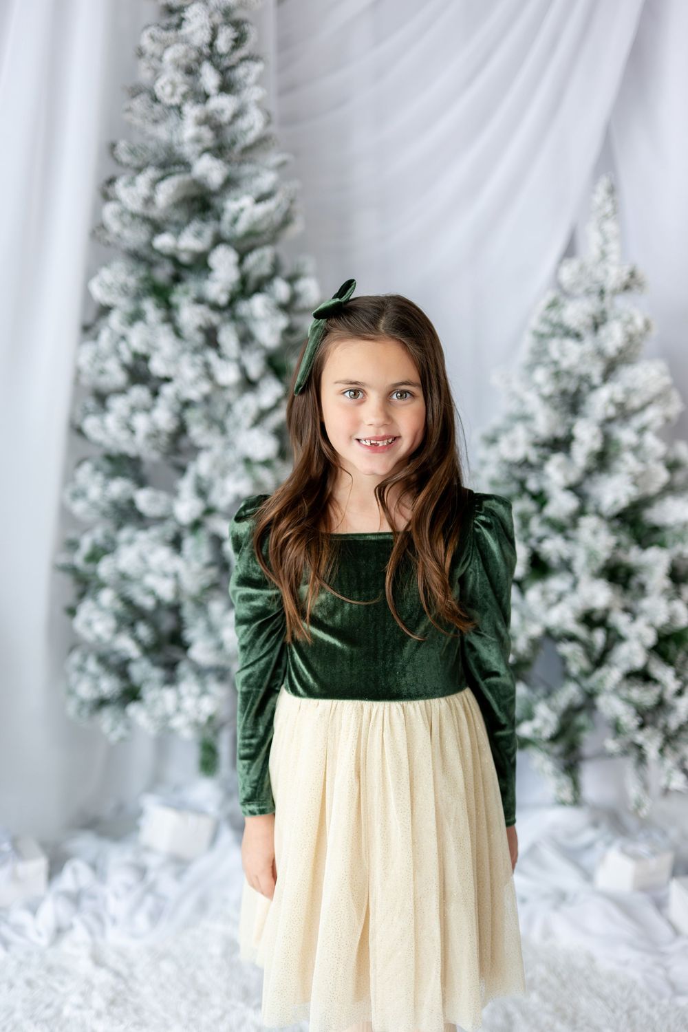 Young girl in green velvet top and cream tulle skirt poses before snow-flocked Christmas trees.