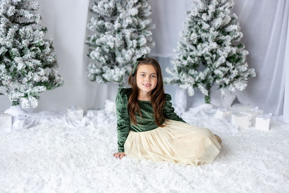 Smiling girl in green velvet and cream tulle dress sits on white fluffy rug by snowy Christmas trees.