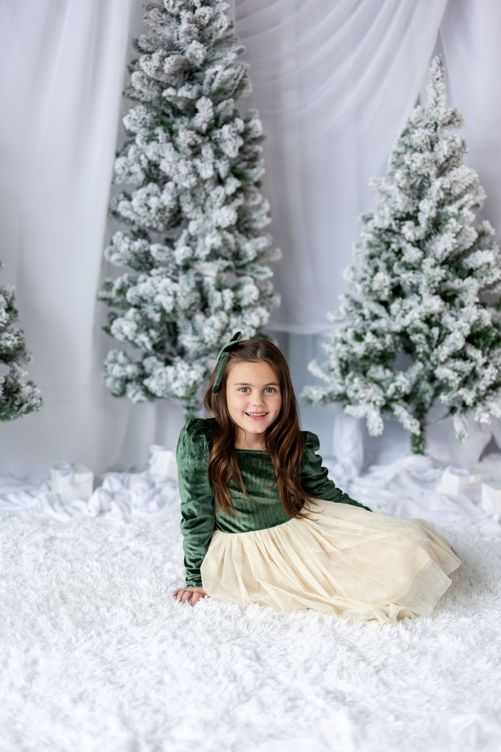 Happy girl in festive green velvet dress crawls playfully on white rug among snow-covered Christmas trees.