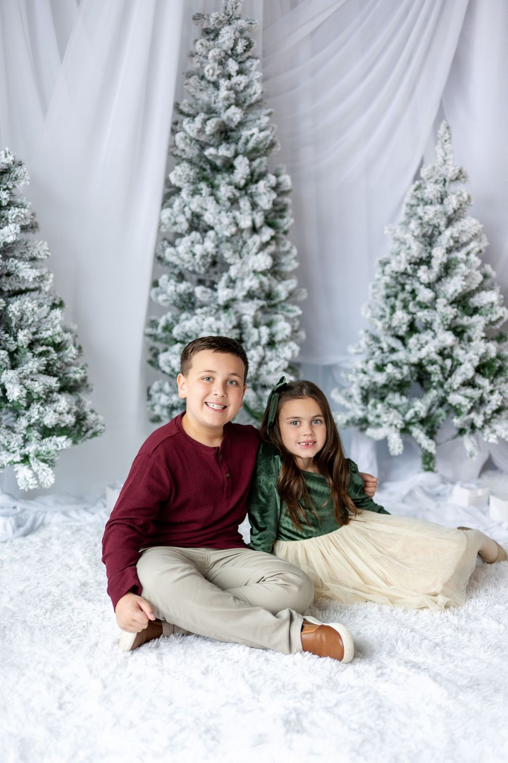 Smiling siblings in holiday outfits sit together on white fluffy rug surrounded by snow-flocked Christmas trees.