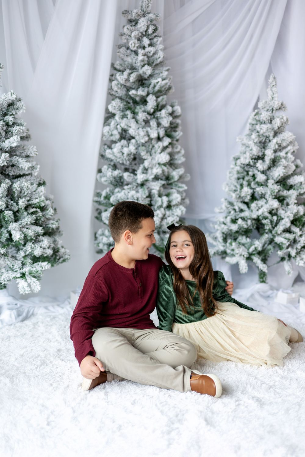 Brother and sister laugh together sitting on white rug surrounded by snow-flocked Christmas trees.