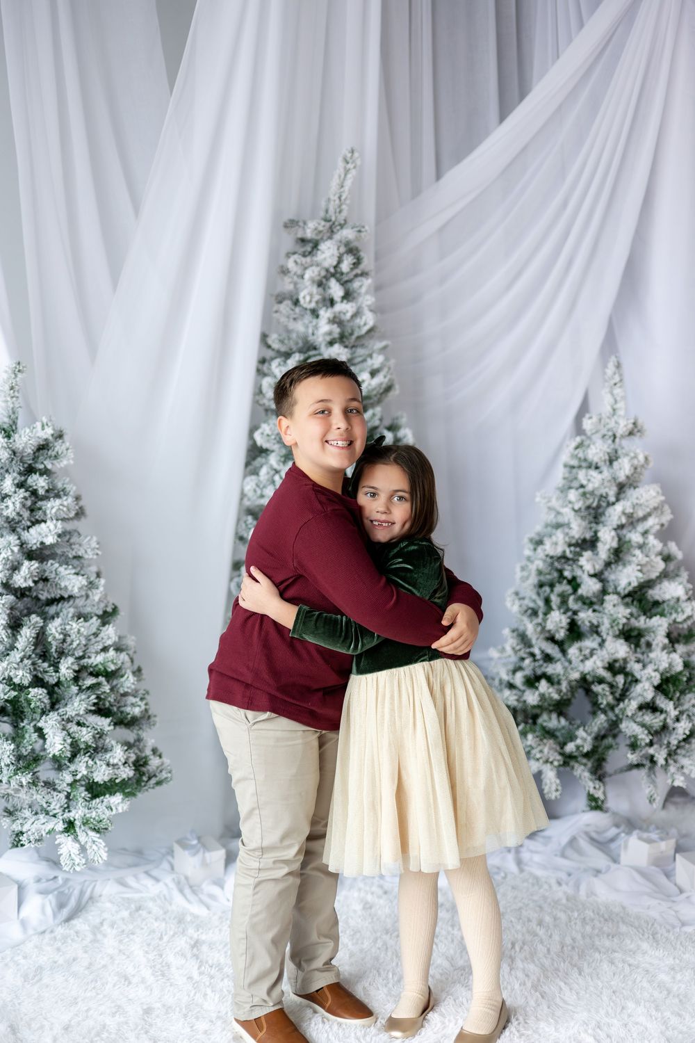 Brother and sister share a warm hug in front of snow-flocked Christmas trees in a white studio setting.