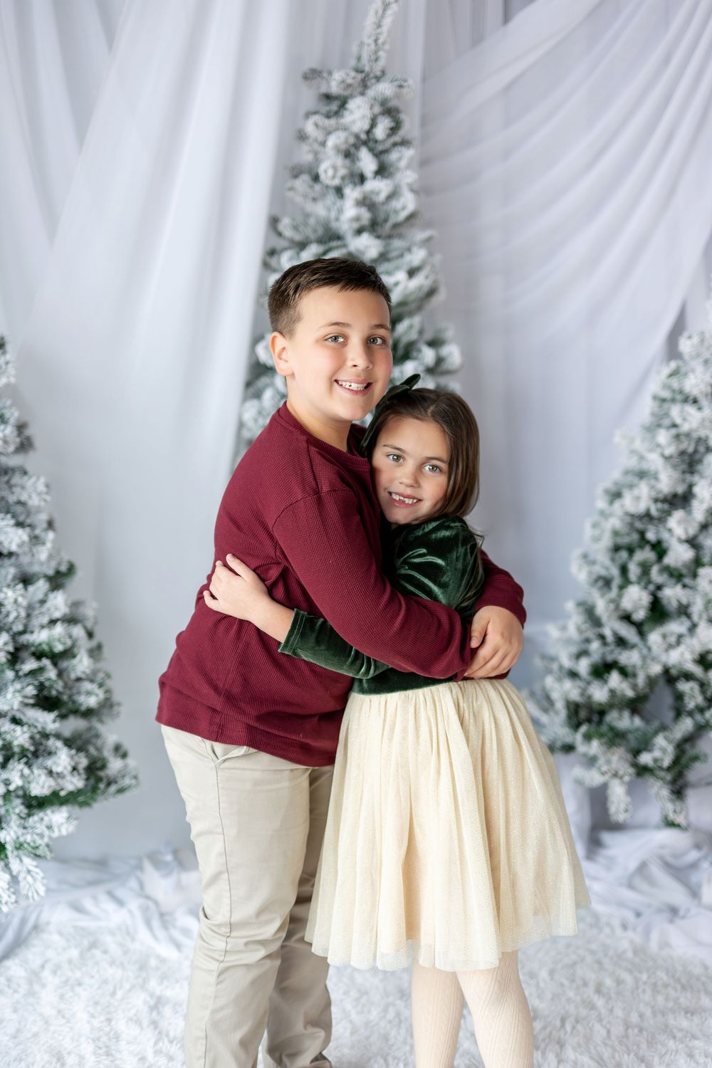 Siblings share a joyful hug in front of snow-flocked Christmas trees in a bright white holiday studio.