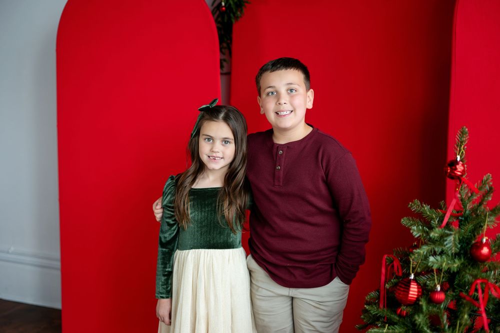 A boy and girl in holiday outfits pose together in front of a red arch with a Christmas tree nearby.