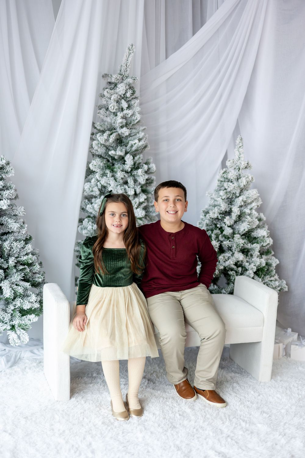 Two children in festive attire sit on a white bench surrounded by snow-flocked Christmas trees.