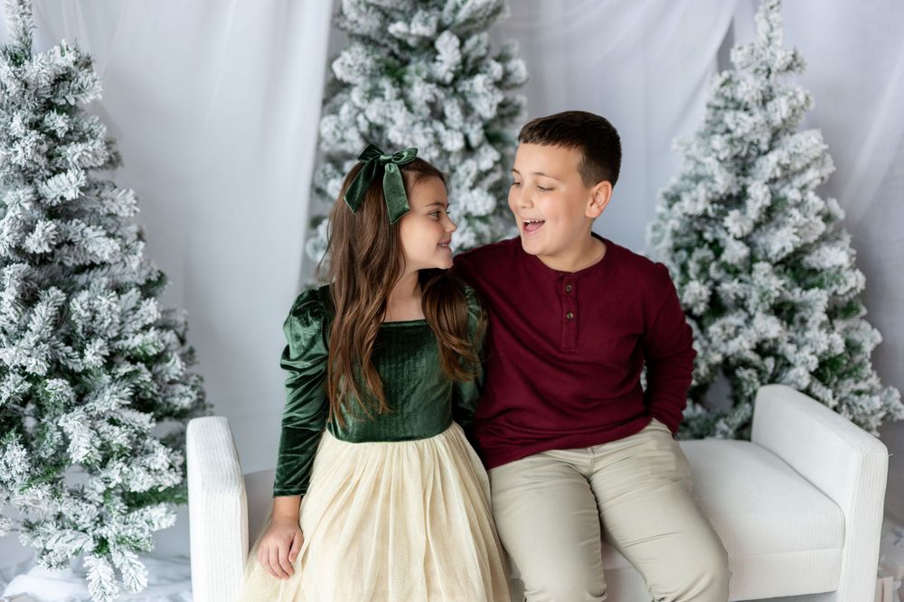 A girl in green velvet dress and boy in burgundy top share a laughing moment by snowy Christmas trees.