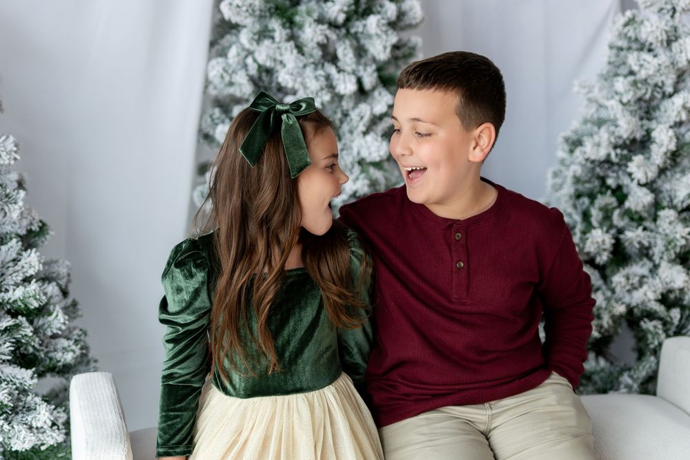 Two children laugh together while sitting near snow-flocked Christmas trees in a white winter setting.