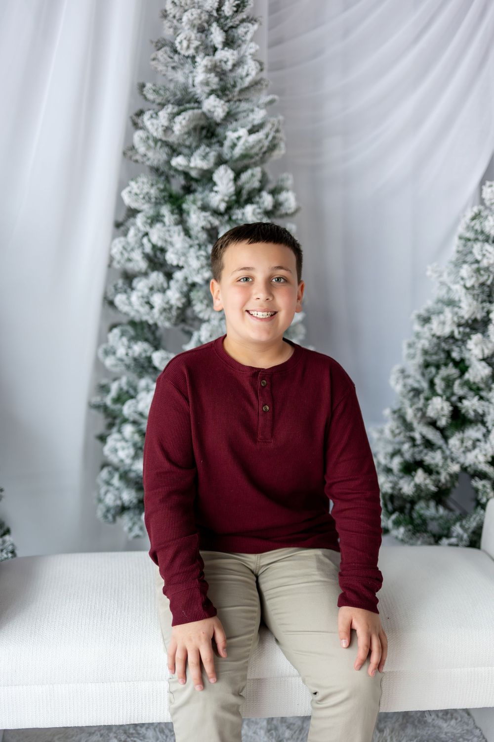 A smiling boy in a burgundy henley sits on a white bench in front of snow-covered Christmas trees.