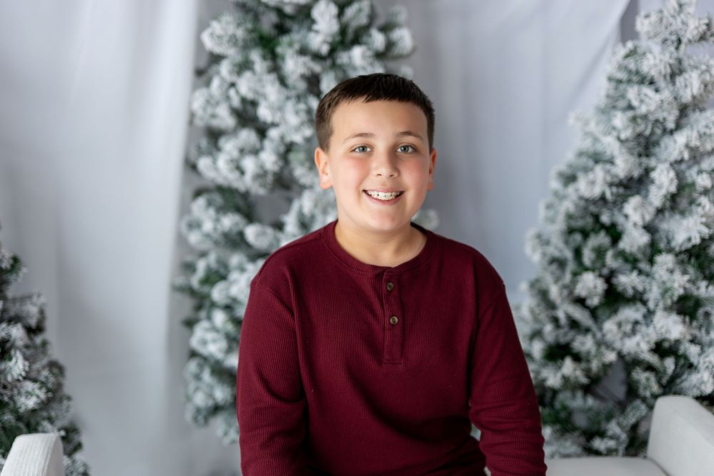 A cheerful boy in a dark red long-sleeve shirt poses in front of snow-flocked holiday trees.