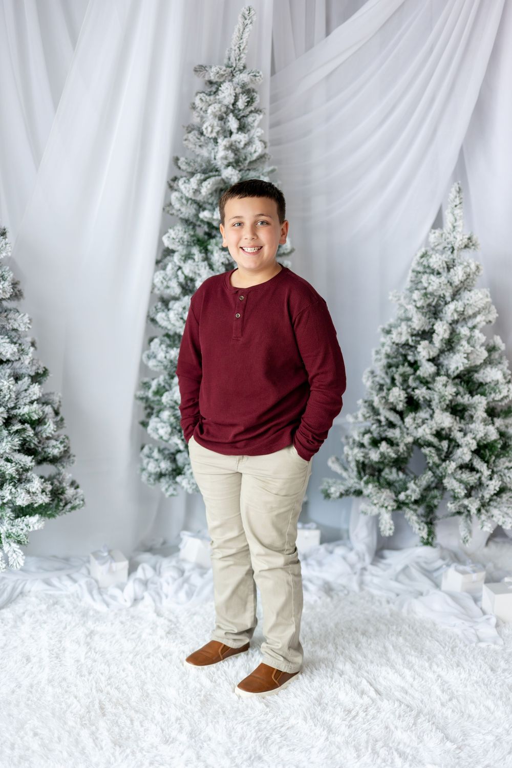 A boy in burgundy top and khaki pants stands smiling among snow-flocked Christmas trees.