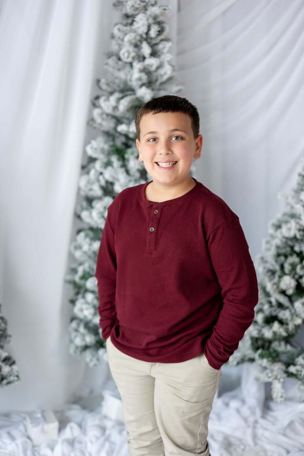 A smiling boy in a maroon henley shirt poses with hands in pockets near snowy Christmas trees.