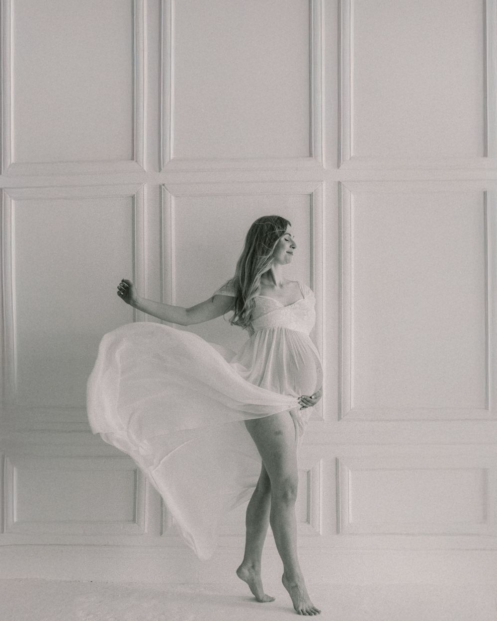 Dynamic black and white maternity portrait with flowing white dress against panelled wall.