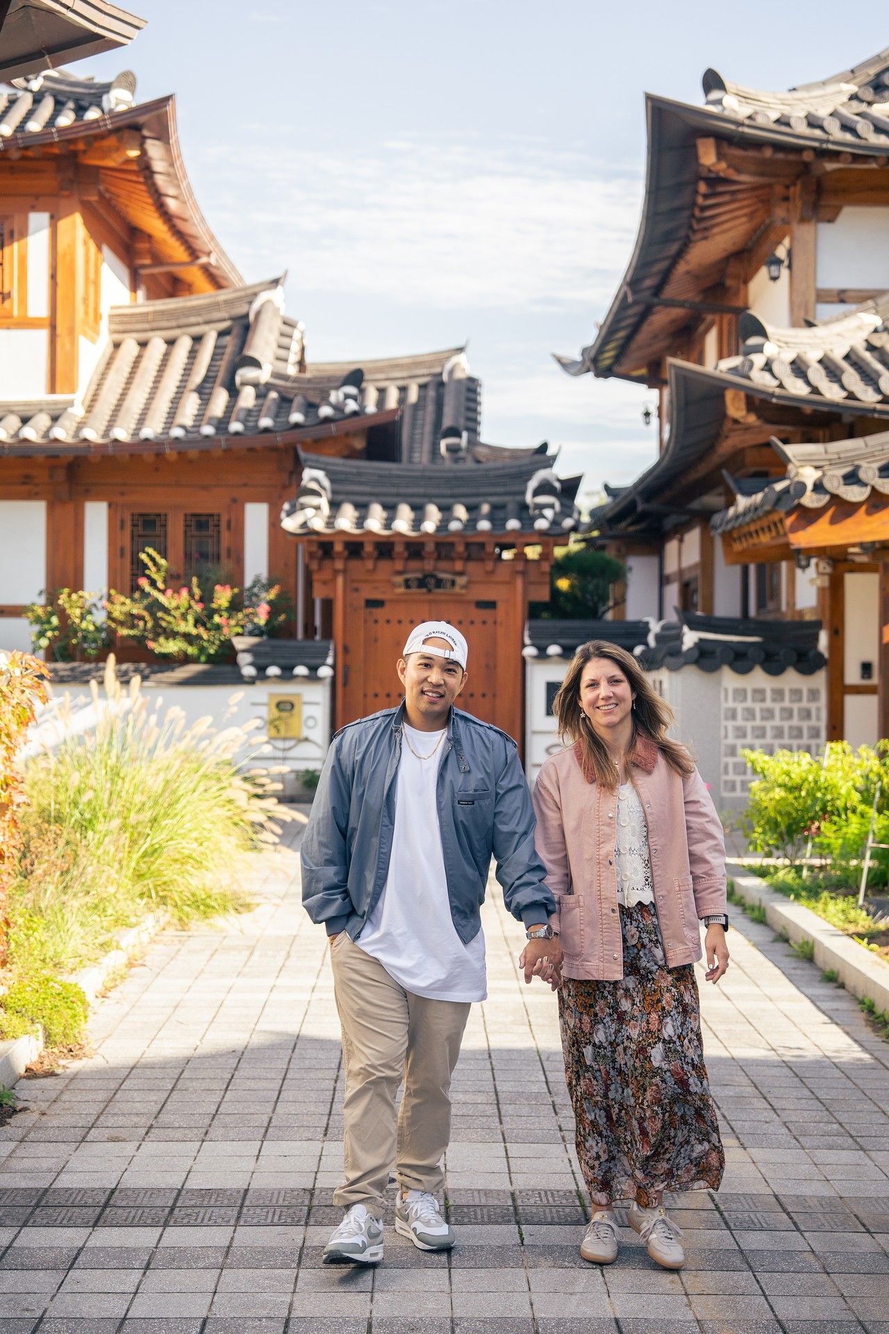 A couple strolls through a traditional Korean hanok village with tiled roofs and wooden architecture in the background.