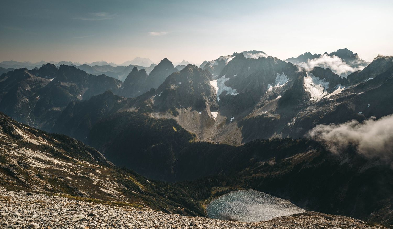 Panoramic view of dramatic mountain range with glaciers, rocky peaks, and low clouds over a valley lake.