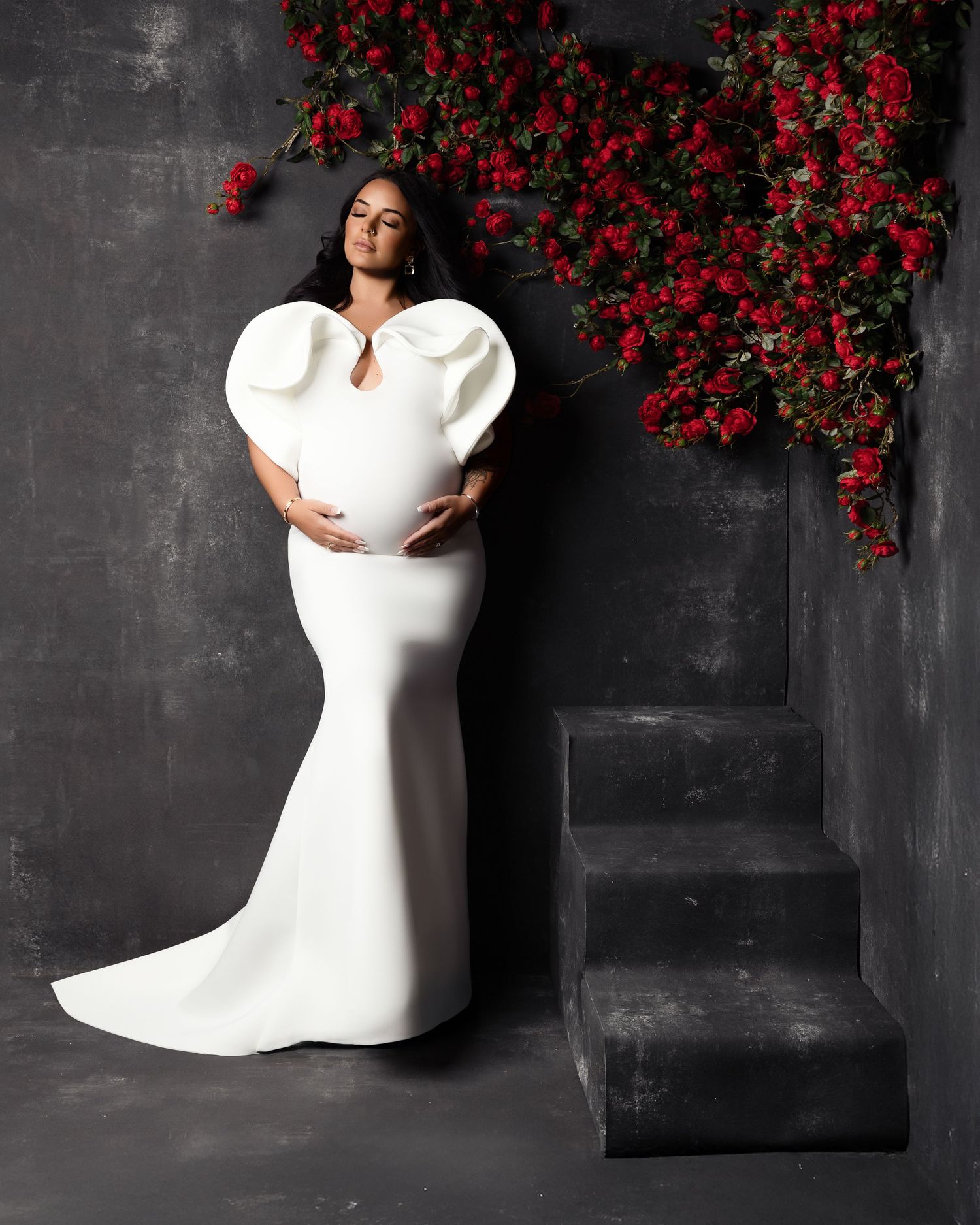 A figure in an elegant white gown with dramatic puff sleeves poses against a dark wall with cascading red flowers.