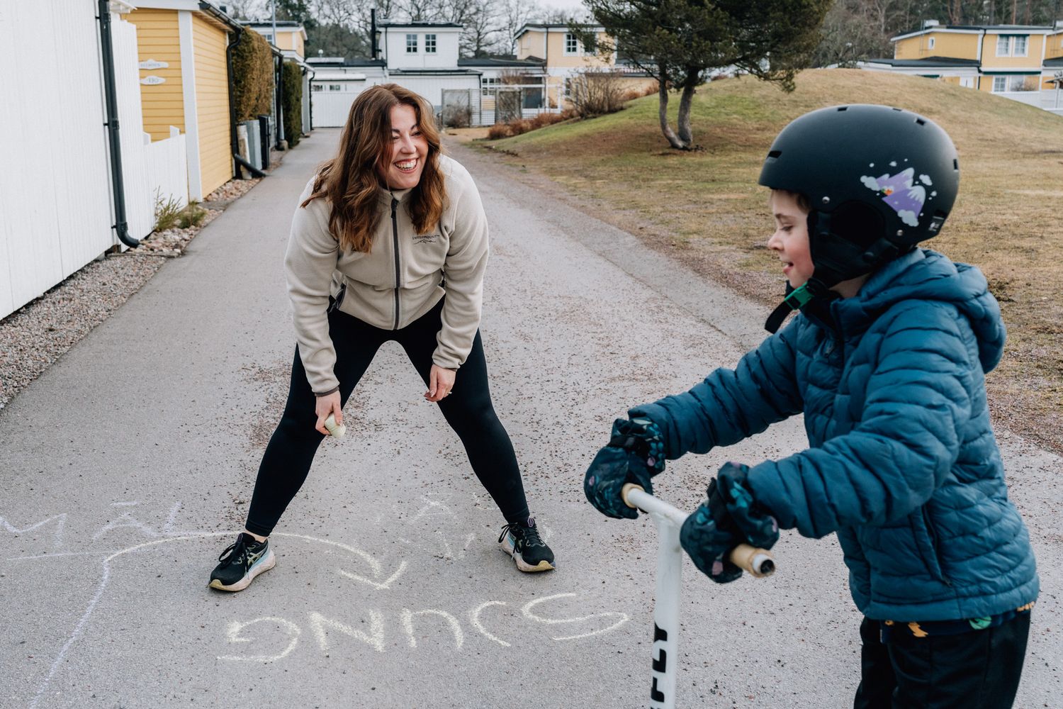 En mamma ritar med gatukritor och tittar på barnet som har en hjälm.