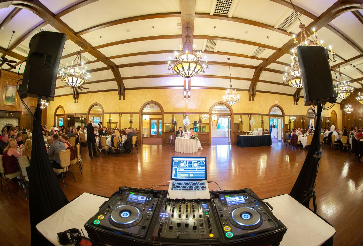 Panoramic view of an elegant ballroom with ornate chandeliers, arched windows, and DJ equipment setup at one end.