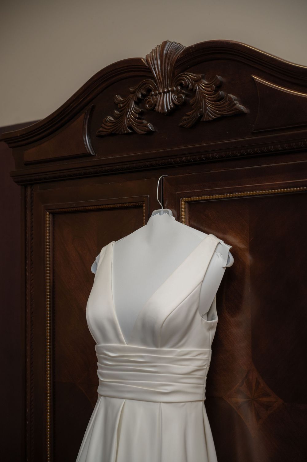 Elegant white wedding dress with V-neck design hanging on ornate wooden armoire.