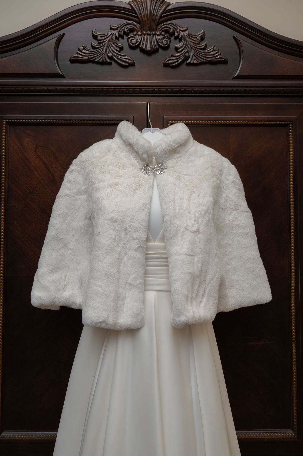 Plush white fur bridal cape with crystal brooch displayed over wedding gown on wooden armoire.