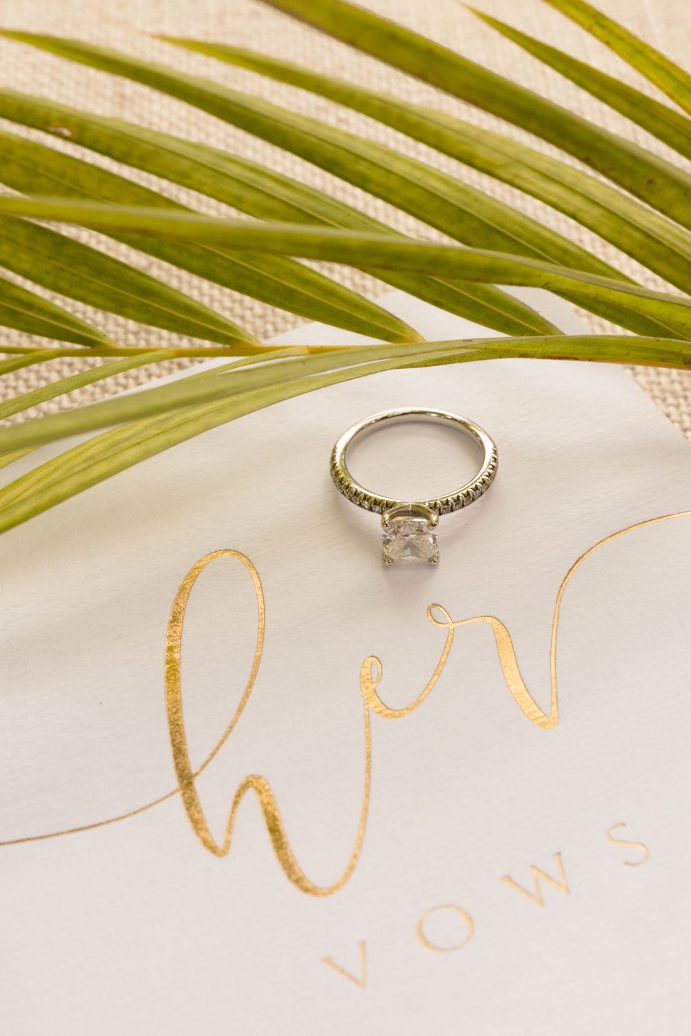 Engagement ring with diamond solitaire displayed on white paper with gold calligraphy and palm frond decor.