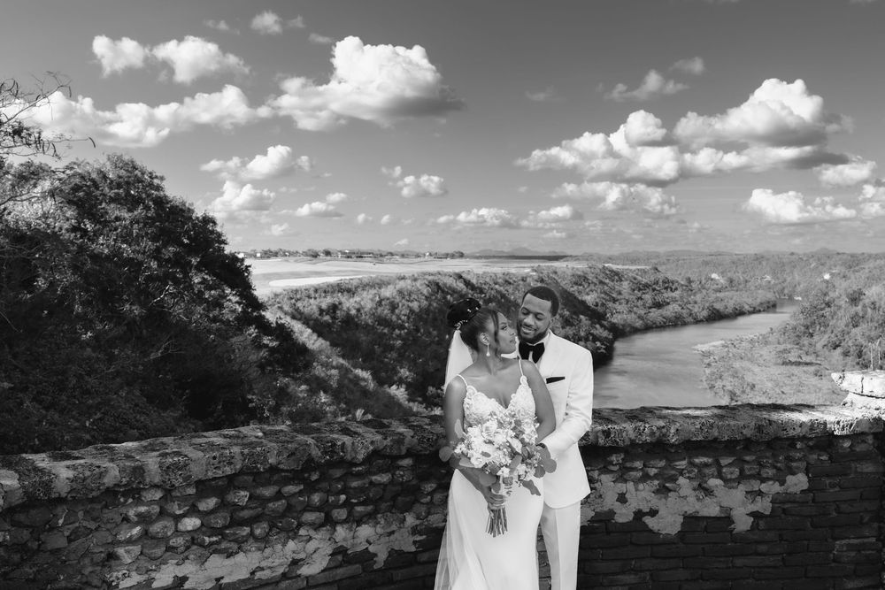A couple shares a romantic moment at a scenic overlook with rolling hills and puffy clouds in black and white.