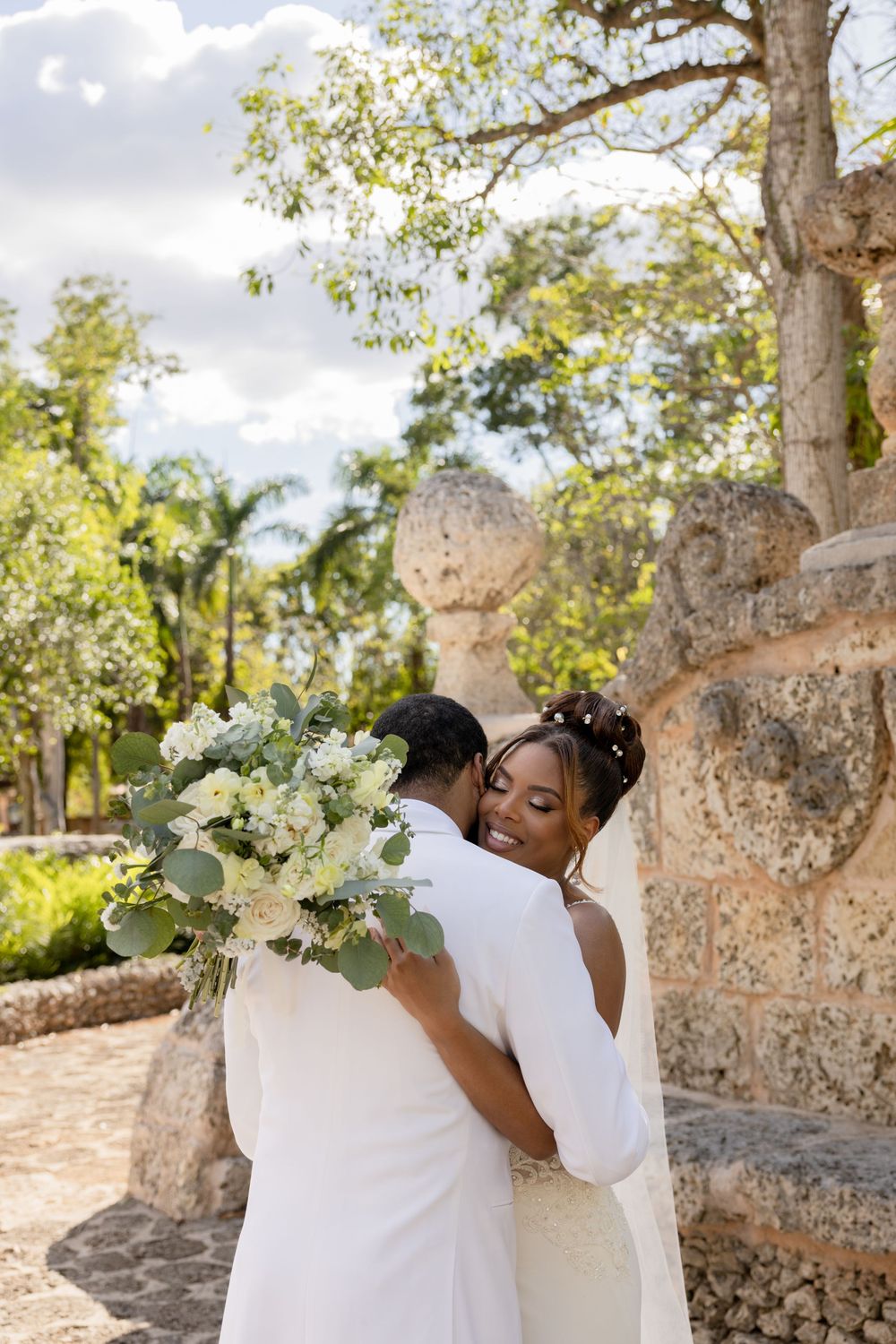 A couple in white wedding attire share an intimate moment among stone walls and trees in a romantic outdoor setting.