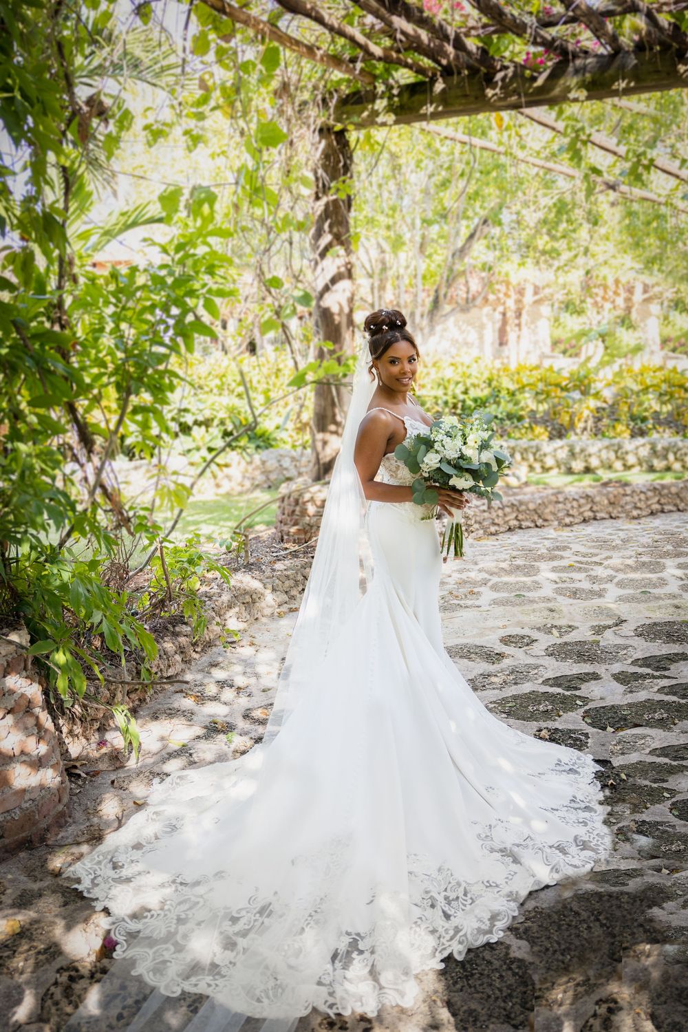 A bride in a flowing white gown poses gracefully on stone steps surrounded by lush greenery and natural light.
