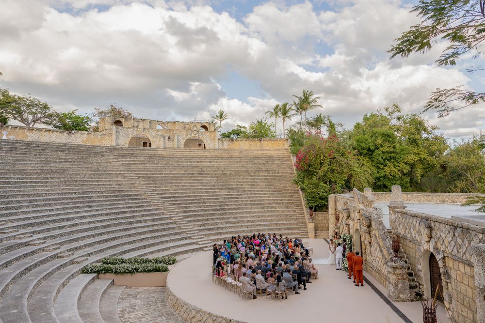 Wedding ceremony at an outdoor amphitheater with stone architecture and palm trees under cloudy skies.