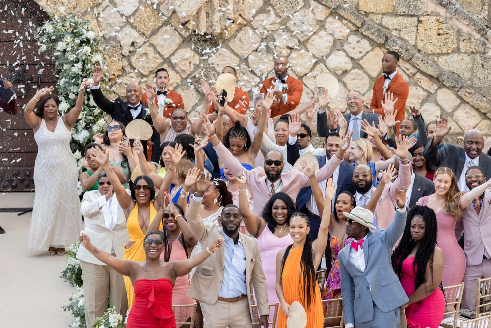 Large group of wedding guests cheering and celebrating together outdoors against a stone wall backdrop.