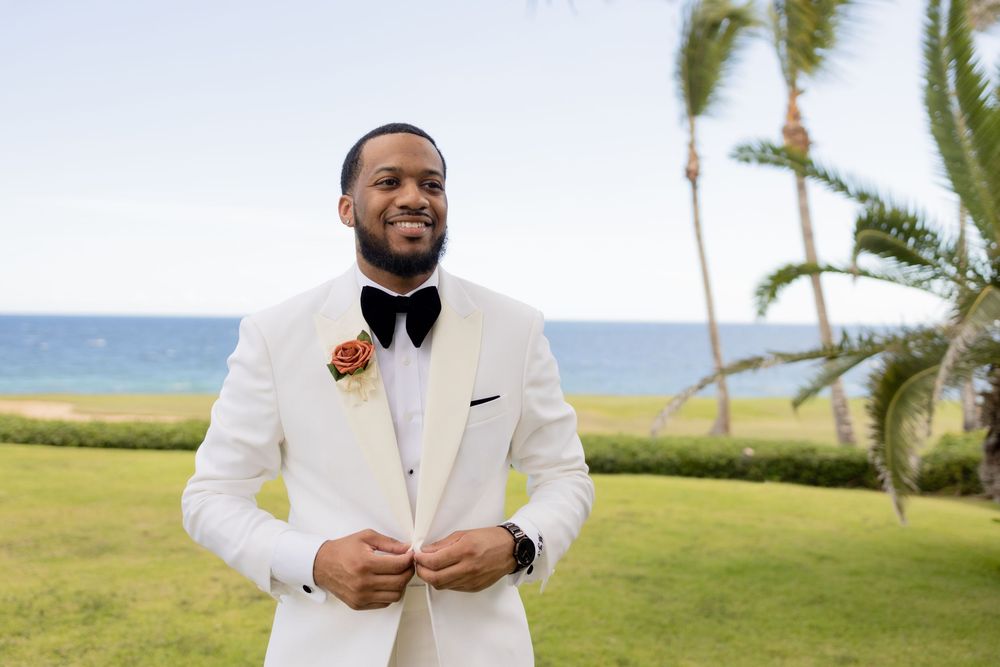 A groom in a white tuxedo poses for wedding photos in a tropical beach setting with palm trees.
