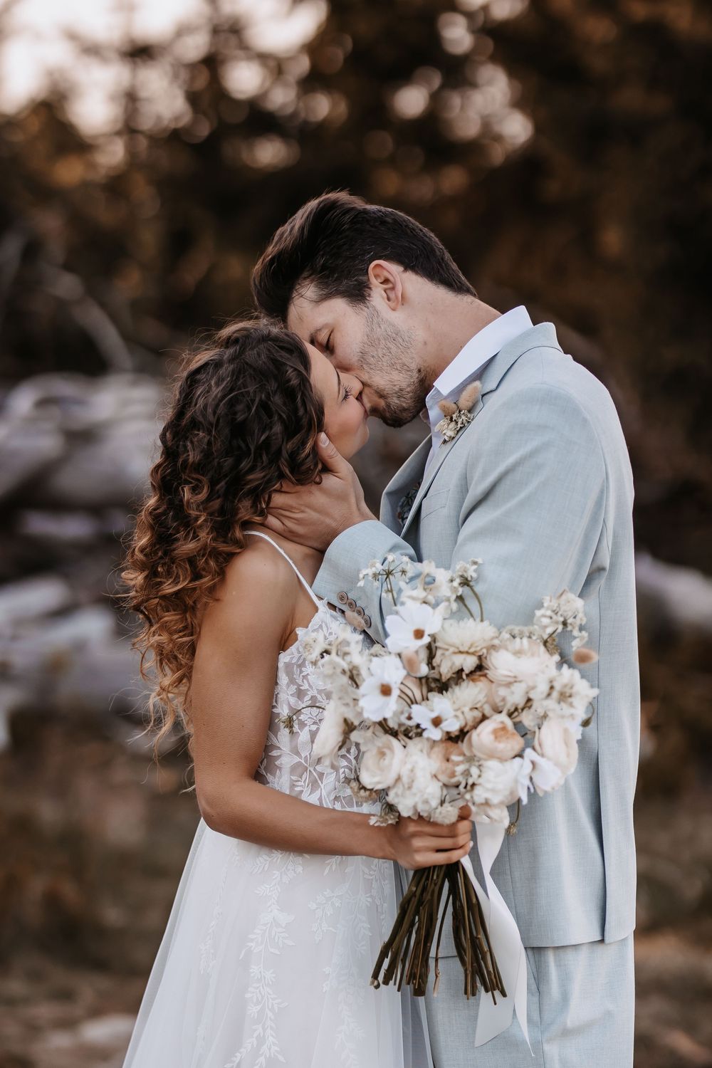 Romantic wedding couple embraces while holding a white floral bouquet against a moody natural backdrop.