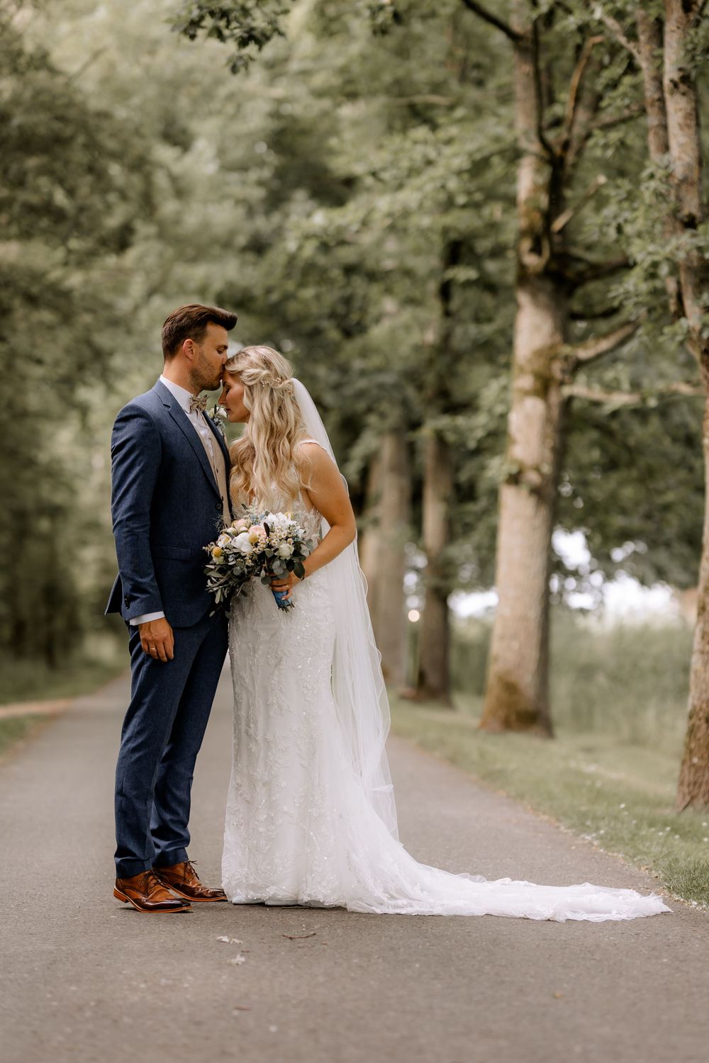 Wedding couple poses on a tree-lined path with the bride's dress trailing behind.