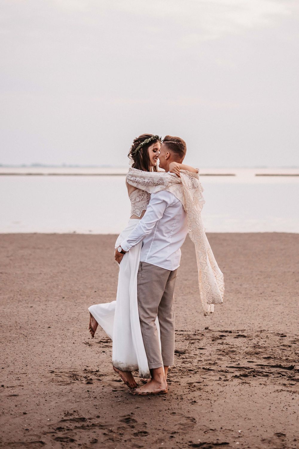 Couple dances and embraces on beach in white and neutral colored outfits.