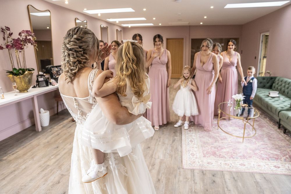 Bride holding a flower girl surrounded by bridesmaids in pink dresses in an elegant bridal suite.