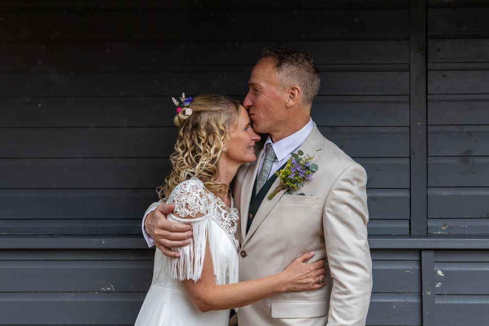 A bride and groom share a kiss outdoors, dressed in wedding attire with floral accessories.