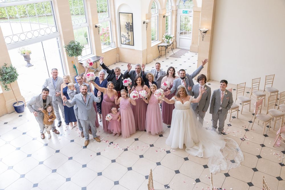 Wedding party members gather in a bright indoor venue wearing pink and gray formal attire for group photos.