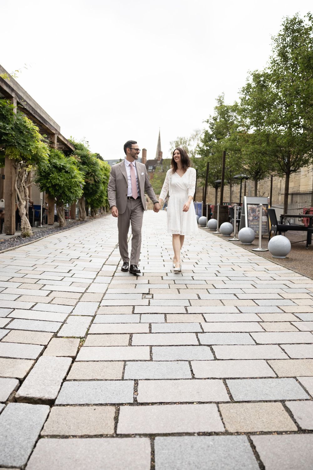 A couple in wedding attire walking together on a stone paved path surrounded by greenery and modern architecture.