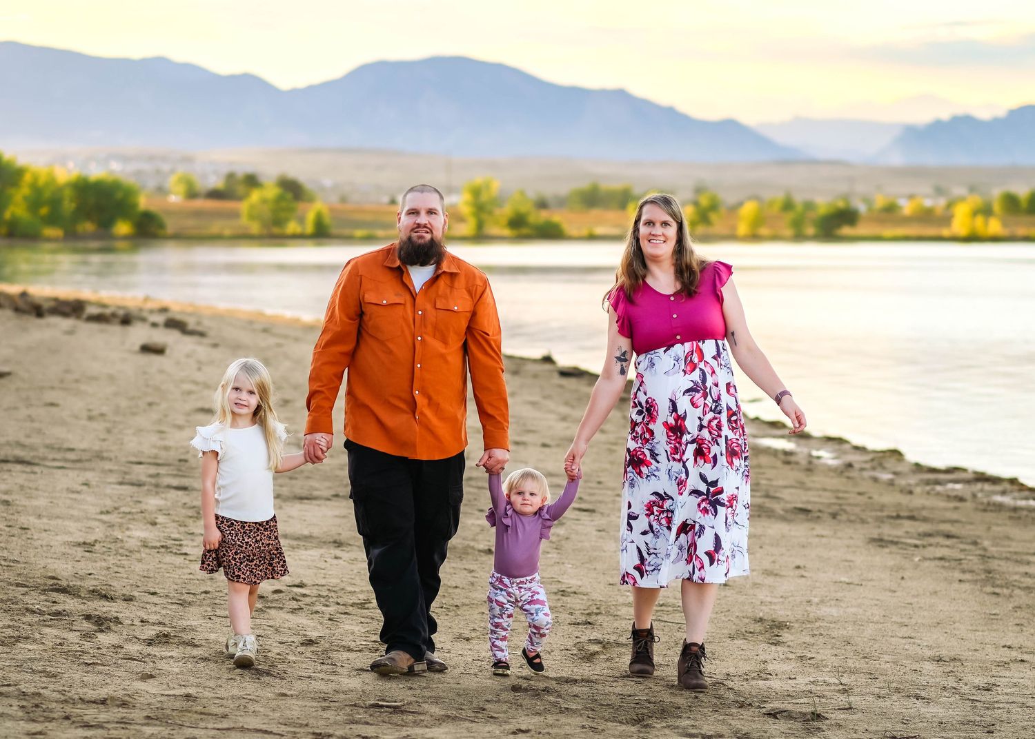 A family walks along a sandy beach shoreline with mountains visible in the distance at sunset.