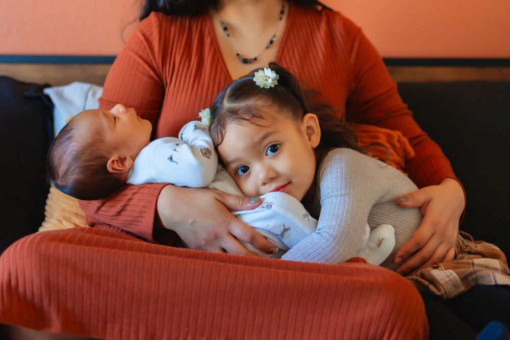 Toddler girl snuggling with mom and newborn baby.  Focus on toddlers face snuggled into babys belly.