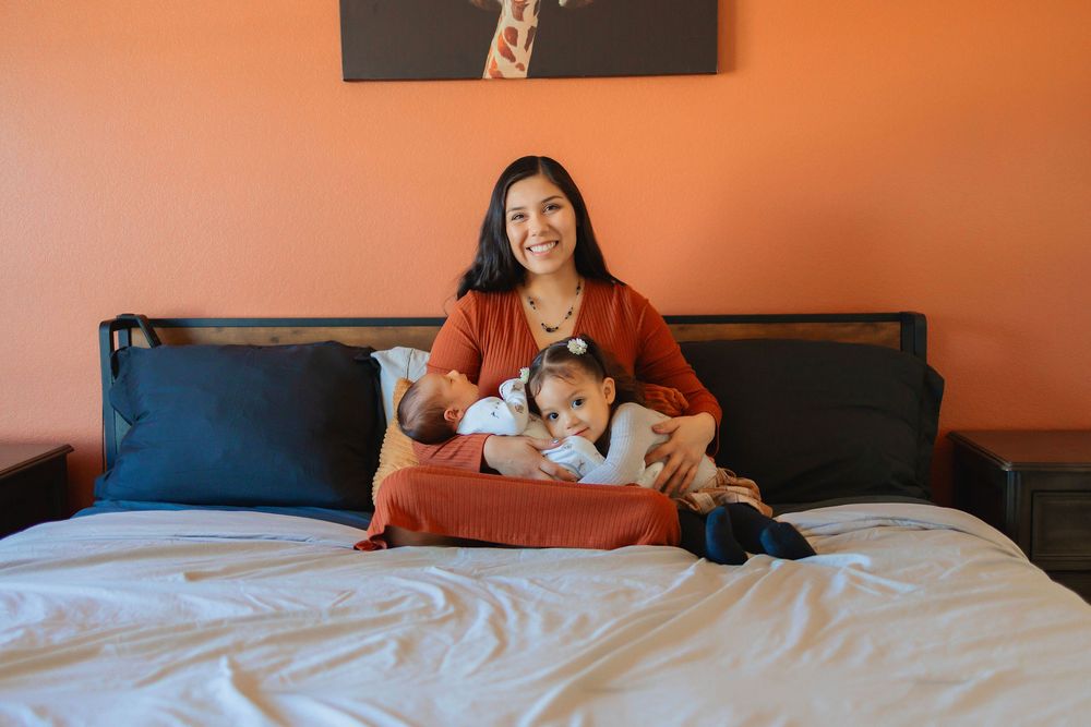 Woman sat on a bed, orange wall, smiling at camera, newborn in her arms, toddler laying on the other side in her arms